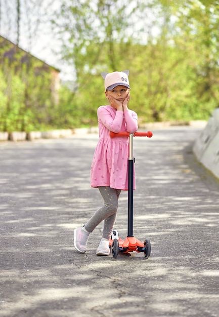 Una niña pensativa con un vestido rojo en un scooter y una gorra en la cabeza