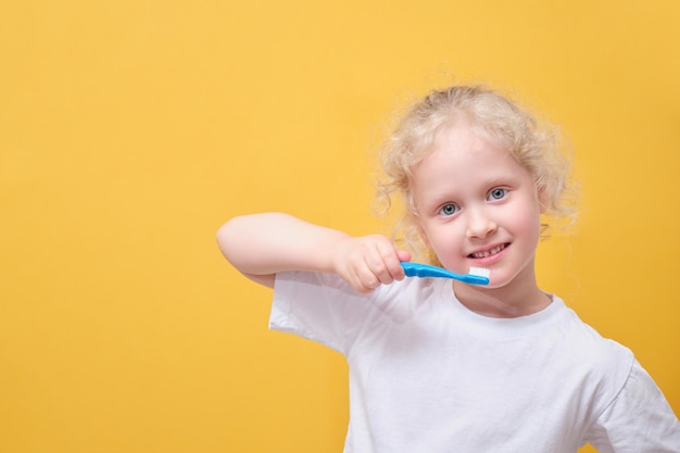 Niña pequeña de 6 años sosteniendo un cepillo de dientes en la mano cepillando el fondo amarillo de los dientes