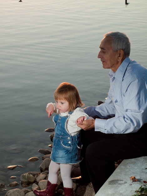 Niña pequeña con abuelo por lago al atardecer.