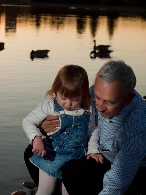 Niña pequeña con abuelo por lago al atardecer.