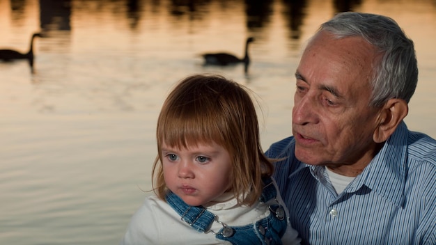 Foto niña pequeña con abuelo por lago al atardecer.