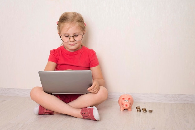 niña pequeña con anteojos usando una computadora portátil sentada en el suelo con una alcancía y monedas