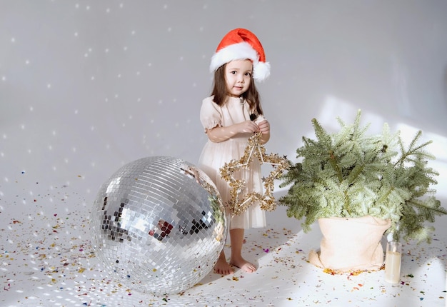 Niña pequeña asiática se para en un sombrero de Papá Noel y sostiene un juguete de Navidad Cerca de la bola de discoteca