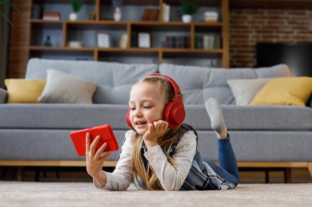 Niña pequeña con auriculares rojos tirados en la alfombra en casa Linda niña sonriente sosteniendo un teléfono inteligente viendo dibujos animados navegando por Internet o viendo videos en las redes sociales jugando juegos en línea