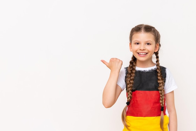 Una niña pequeña con una bandera alemana en una camiseta con el pelo largo en coletas señala con el dedo índice de lado a su anuncio en un fondo blanco aislado Cursos de idiomas Copiar espacio