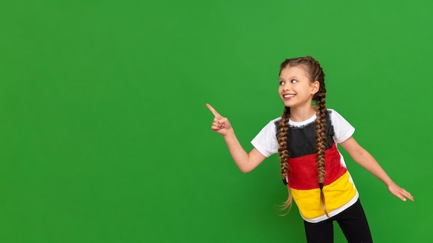 Una niña pequeña con una bandera alemana en su camiseta señala su anuncio en un fondo verde aislado Cursos de alemán para escolares Copiar espacio