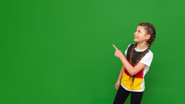 Una niña pequeña con una bandera alemana en su camiseta señala su anuncio en un fondo verde aislado Cursos de alemán para escolares Copiar espacio