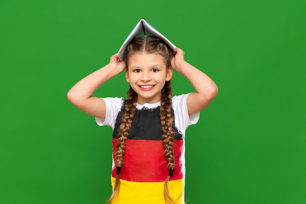 Una niña pequeña con la bandera de Alemania en su camiseta sostiene cuadernos sobre su cabeza como una casa en un fondo verde aislado