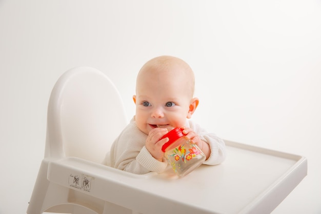 La niña pequeña bebe agua de una botella en el interior se sienta en una silla alta