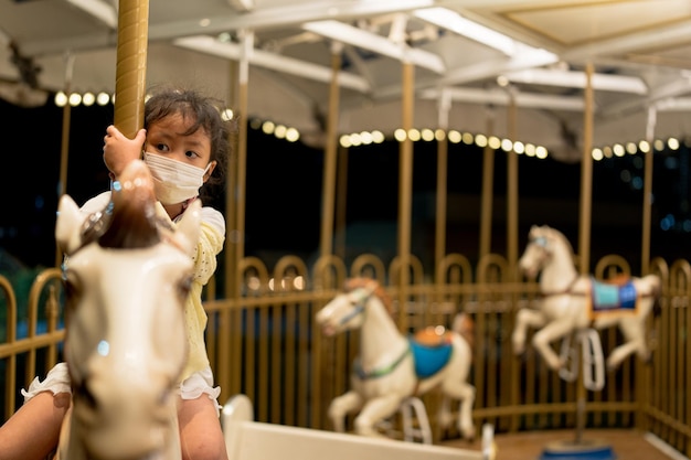 Foto niña pequeña en caballo de carrusel con mascarilla saludable para proteger el virus y la contaminación.