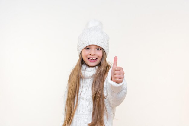 Una niña pequeña con un cálido sombrero blanco y un suéter de punto muestra su pulgar hacia arriba y sonríe. fondo aislado.