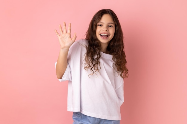 Niña pequeña con camiseta blanca sonríe con dientes y levanta la palma de la mano saluda a un amigo diciendo hola o adiós