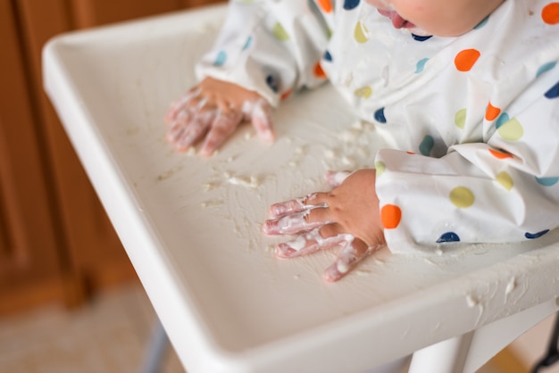 Una niña pequeña en una camiseta y un plato sentado en la silla de un niño comiendo con las manos cereales con yogur comida