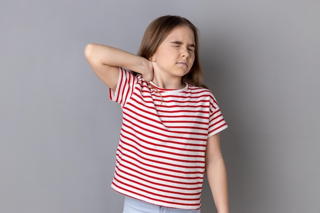 Foto niña pequeña con camiseta a rayas de pie masajeando el cuello para aliviar el dolor de la tensión muscular en la espalda