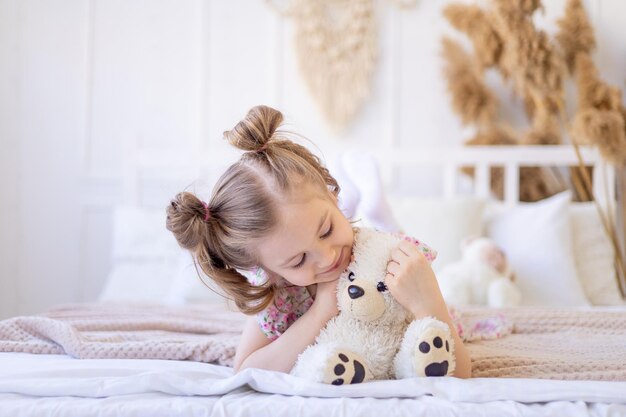 Una niña pequeña en casa en un tranquilo interior natural de color beige juega y se abraza con un suave oso de peluche