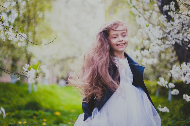 Niña pequeña con chaqueta negra y vestido blanco corriendo en el jardín de cerezos de primavera