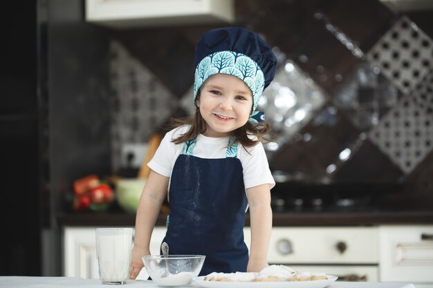 Una niña pequeña en la cocina con un delantal y un sombrero de chef rocía galletas con azúcar en polvo