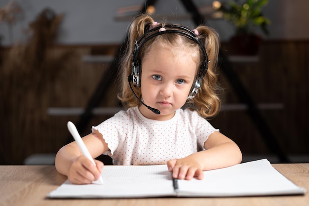 Una niña pequeña, una colegiala, se sienta en casa en la mesa sola por la noche cerca de un cuaderno. Sostiene un bolígrafo y escribe algo.