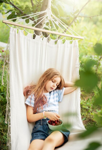 Una niña pequeña descansa en una hamaca y come cerezas en el verano