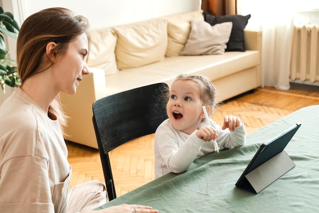 Una niña pequeña descubrió cómo hacer tareas en una tableta en casa o completar un juego Idea de iluminación Mamá ayuda a hacer la tarea para el jardín de infantes de su hija