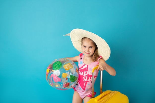 Niña pequeña y divertida con pelota de playa sosteniendo una maleta amarilla sobre fondo azul Verano