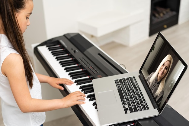 Niña pequeña en la escuela en el hogar aprendiendo piano desde la computadora que se conecta a la clase en línea de música en Internet por el maestro de la escuela. Nuevo estilo de vida normal y educación, estudio de estudiantes en el concepto de hogar.