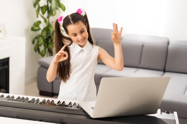 Niña pequeña en la escuela en el hogar aprendiendo piano desde la computadora que se conecta a la clase en línea de música en Internet por el maestro de la escuela. Nuevo estilo de vida normal y educación, estudio de estudiantes en el concepto de hogar.