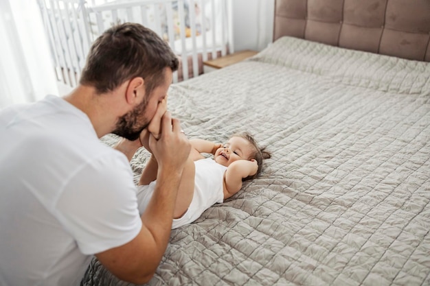 Una niña pequeña está acostada en la cama en el dormitorio y se ríe mientras su padre juega con sus pies.