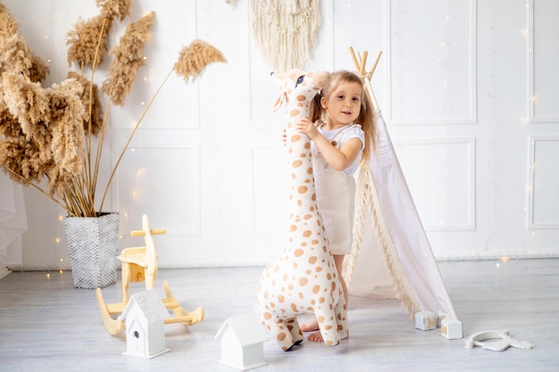 Una niña pequeña está jugando con una jirafa grande tan alta como ella en casa en una guardería luminosa y riendo