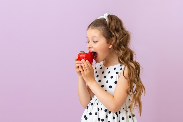 Una niña pequeña está a punto de morder un trozo de pimienta sobre un fondo rosa aislado Una hermosa niña con cabello rizado y un vestido de lunares blancos