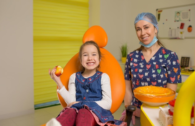 Una niña pequeña está sentada en una silla dental naranja sonriendo con una manzana roja en sus manos Pediátrica