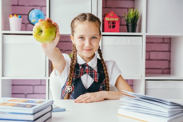 Foto niña pequeña una estudiante de escuela se sienta en su escritorio y extiende una manzana