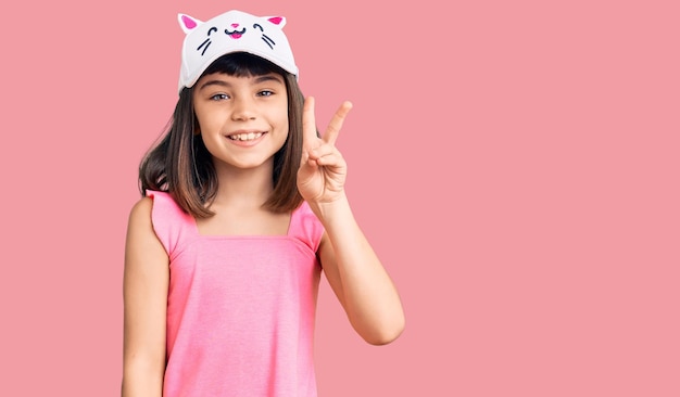 Niña pequeña con una explosión usando una gorra de gatito divertida sonriendo con cara feliz guiñando un ojo a la cámara haciendo el signo de la victoria. número dos.