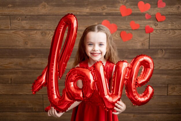 Una niña pequeña feliz en pijama rojo con un gran regalo en sus manos sobre un fondo de madera marrón oscuro se ríe y sonríe el concepto del día de San Valentín un lugar para el texto