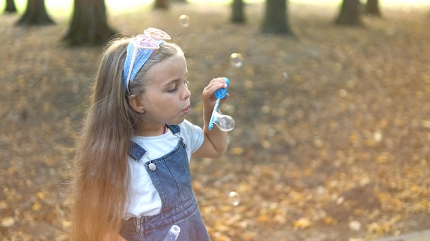 Niña pequeña feliz que sopla pompas de jabón afuera en un parque verde. Concepto de actividades de verano al aire libre.