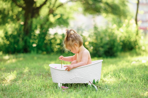 Niña pequeña feliz toma un baño de leche con pétalos. Niña en un baño de leche en un verde. Ramos de peonías rosas. Bebé bañándose. Higiene y cuidado de niños pequeños.