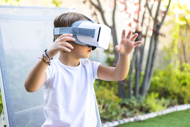 Niña pequeña con gafas de realidad virtual mirando a la derecha tratando de tocar algo de forma virtual sentada en una tumbona en el jardín de su casa Concepto digital del juego Metaverse VR