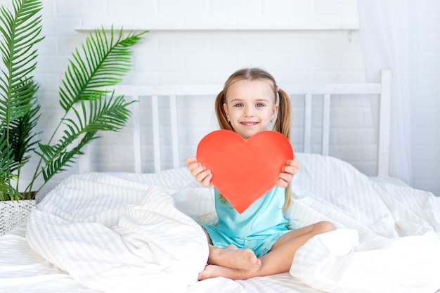 Niña pequeña con un gran corazón rojo en las manos sentada en casa en la cama en una cama de algodón blanco y sonriendo dulcemente el concepto del día de san valentín