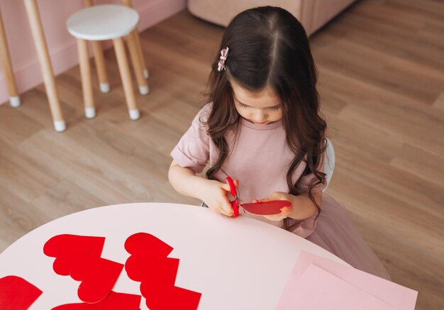 Una niña pequeña hace tarjetas de San Valentín con tijeras de papel de colores y lápiz sentada en una mesa