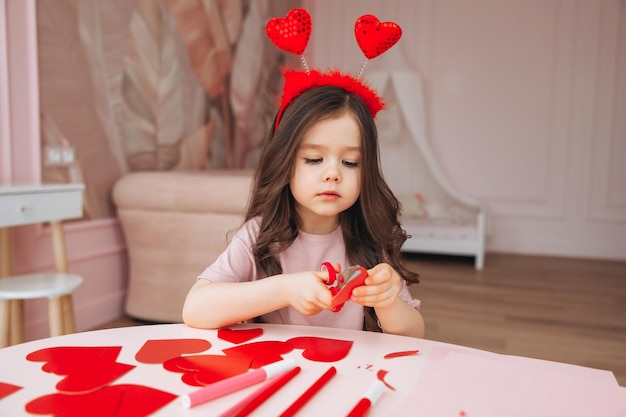 Una niña pequeña hace tarjetas de San Valentín con tijeras de papel de colores y lápiz sentada en una mesa