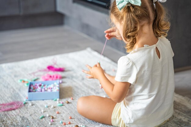 Foto niña pequeña haciendo una pulsera de cuentas de madera en casa sala de estar la creatividad de los niños y el desarrollo de las habilidades motoras finas