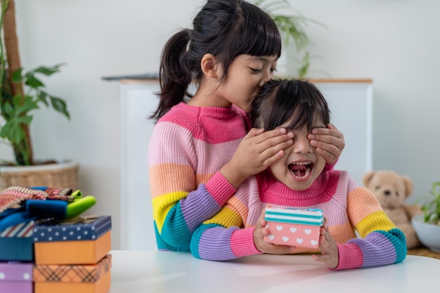 Niña pequeña haciendo regalos a una niña feliz sosteniendo una caja de regalo, cerrando los ojos de una linda sonrisa emocionada con la sorpresa de cumpleaños de su hermana