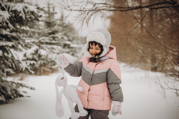 Una niña pequeña con una liebre tejida se para en la calle bajo la nieve voladora. día de invierno