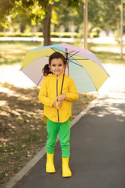 Una niña pequeña y linda con un impermeable amarillo y un paraguas arcoíris multicolor se ríe