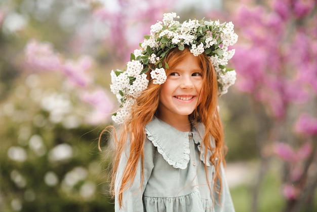 Una niña pequeña y linda que se ríe de 3 a 4 años usa una corona floral y un elegante vestido rústico sobre la naturaleza