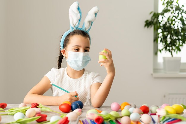 Niña pequeña con una máscara médica pinta huevos de Pascua con un patrón. Quedarse en casa.