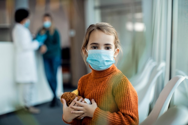 Niña pequeña con mascarilla sosteniendo su peluche mientras está sentada en la sala de espera del hospital