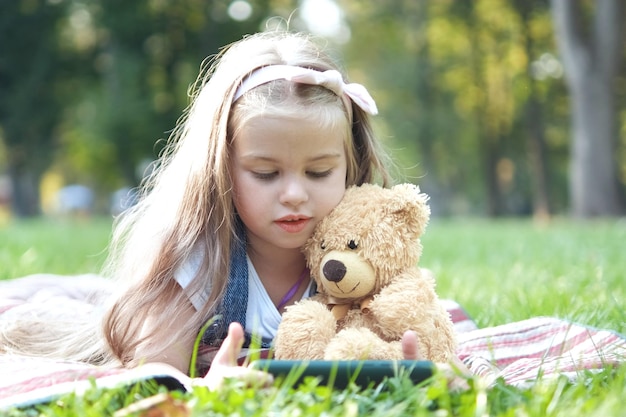 Niña pequeña mirando en su teléfono móvil junto con su osito de peluche favorito al aire libre en el parque de verano