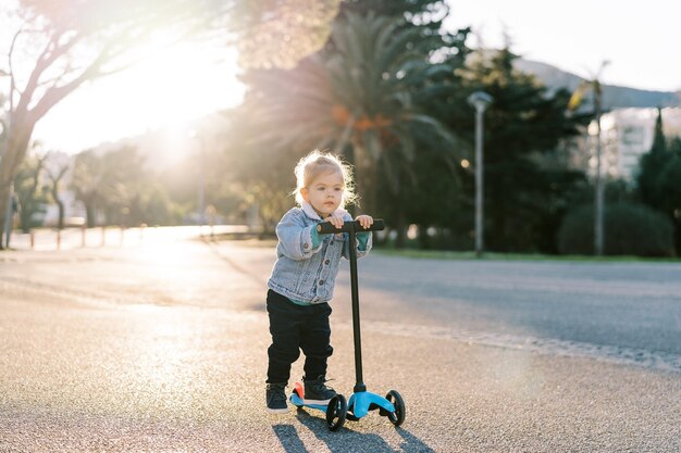 Foto niña pequeña monta un scooter empujando su pie a lo largo de la carretera en un parque soleado