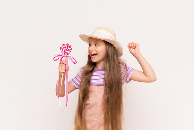 Una niña pequeña muestra una piruleta y sonríe Un niño con un vestido de verano y un sombrero de verano sobre un fondo blanco aislado